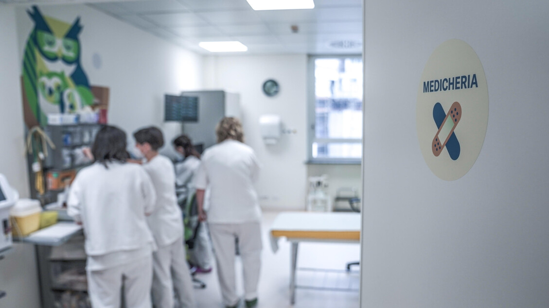 L'immagine mostra un reparto ospedaliero, con un gruppo di persone in una stanza. Tra di loro, si riconosce una persona che indossa pantaloni bianchi e un'altra figura sfocata con un camice bianco. L'ambiente &egrave; caratterizzato da elementi architettonici tipici di un ospedale, con attrezzature mediche e mobili presenti.