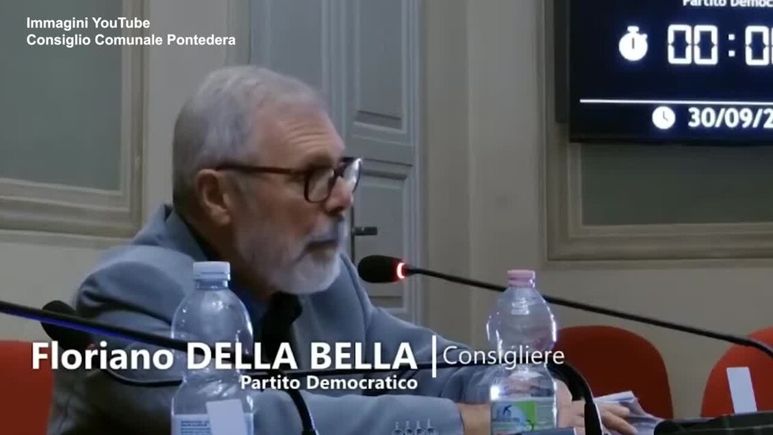 A Pontedera, un uomo, identificato come Floriano Della Bella, &egrave; seduto a un microfono durante una conferenza stampa. Indossa un abito e una cravatta. &Egrave; un consigliere del Partito Democratico e sta parlando in un contesto ufficiale. La scena include altre persone presenti, suggerendo un evento pubblico.