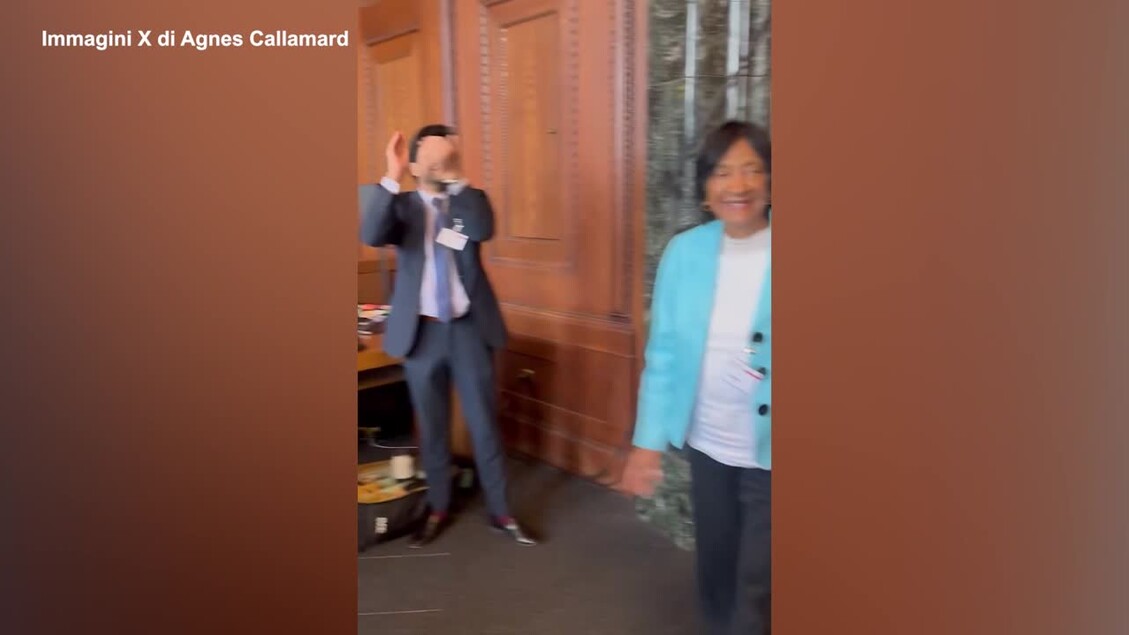 Un uomo in abito formale scherza dicendo: Se applaudite ancora un po', forse lo divider&ograve; con Trump. Una donna &egrave; presente nella stessa stanza. Entrambi mostrano espressioni facciali che possono suggerire sorpresa o felicit&agrave;. L'ambiente &egrave; interno e decorato, con pareti visibili.