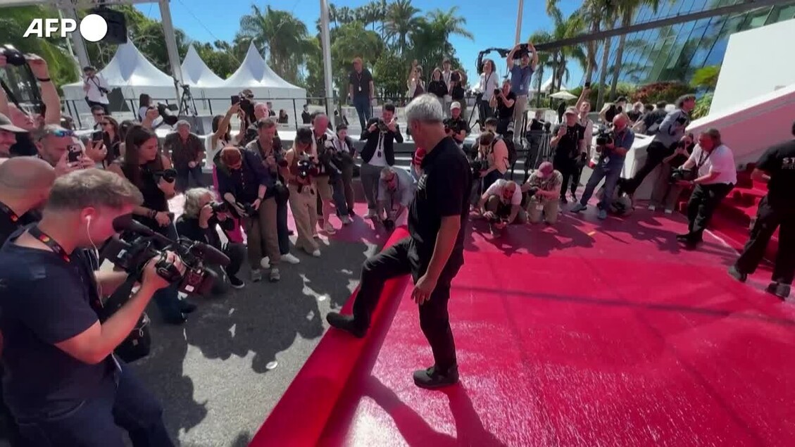 Fervono i preparativi per il 79esimo Festival di Cannes, srotolato il tappeto rosso. Un uomo cammina su questa superficie rossa, mentre una folla di persone osserva. L'evento &egrave; caratterizzato dalla presenza di paparazzi e da un'atmosfera di moda.