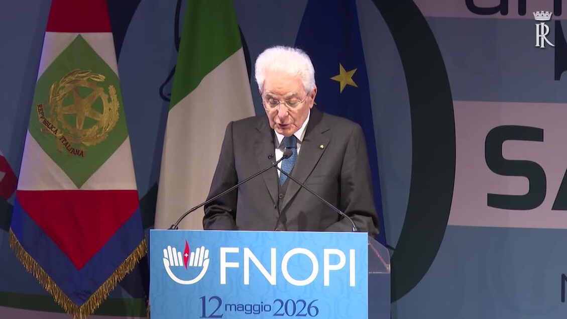 Sergio Mattarella &egrave; riconoscibile nell'immagine mentre parla da un podio. Indossa un abito formale e una cravatta. &Egrave; presente una folla di persone. Una bandiera con una stella dorata &egrave; visibile. Mattarella afferma: Qualcuno cerca di derubricare Covid a leggera influenza.