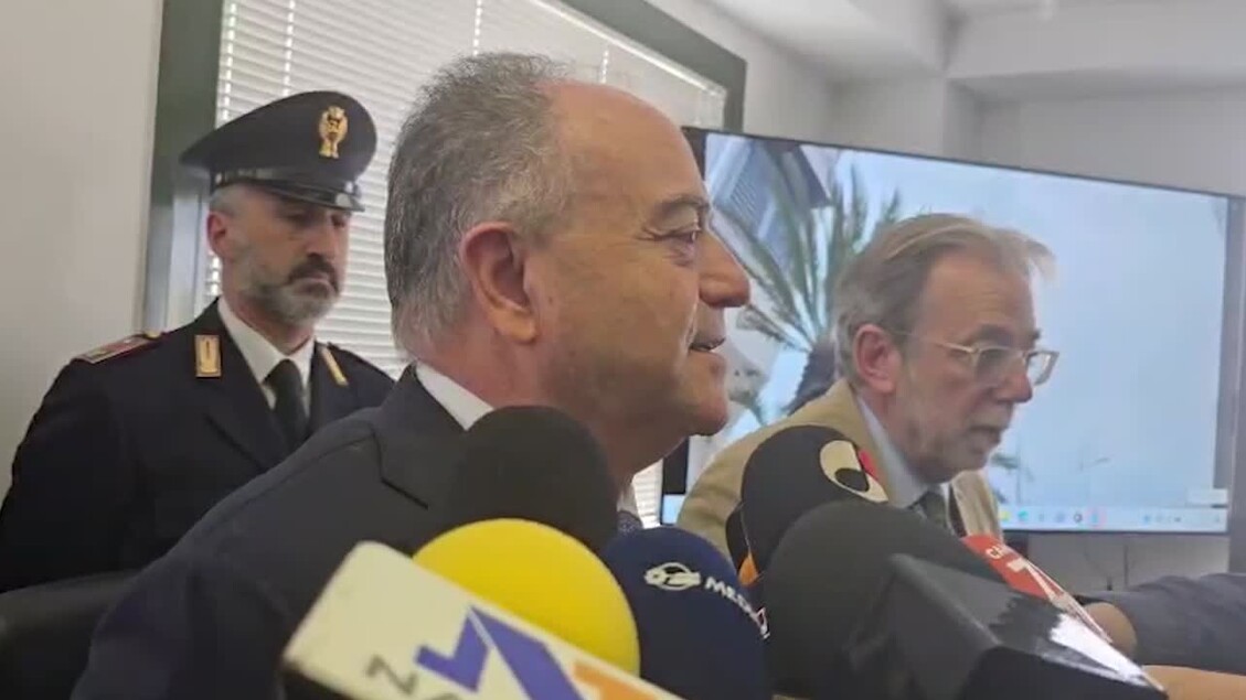 Durante una conferenza stampa, un gruppo di uomini in abiti formali parla in microfoni. Tra di loro, ci sono uomini in uniforme e in giacca. Gratteri afferma: Rubati e venduti da poliziotti infedeli dati su calciatori e cantanti. La scena include anche una folla di persone, suggerendo un evento ufficiale.