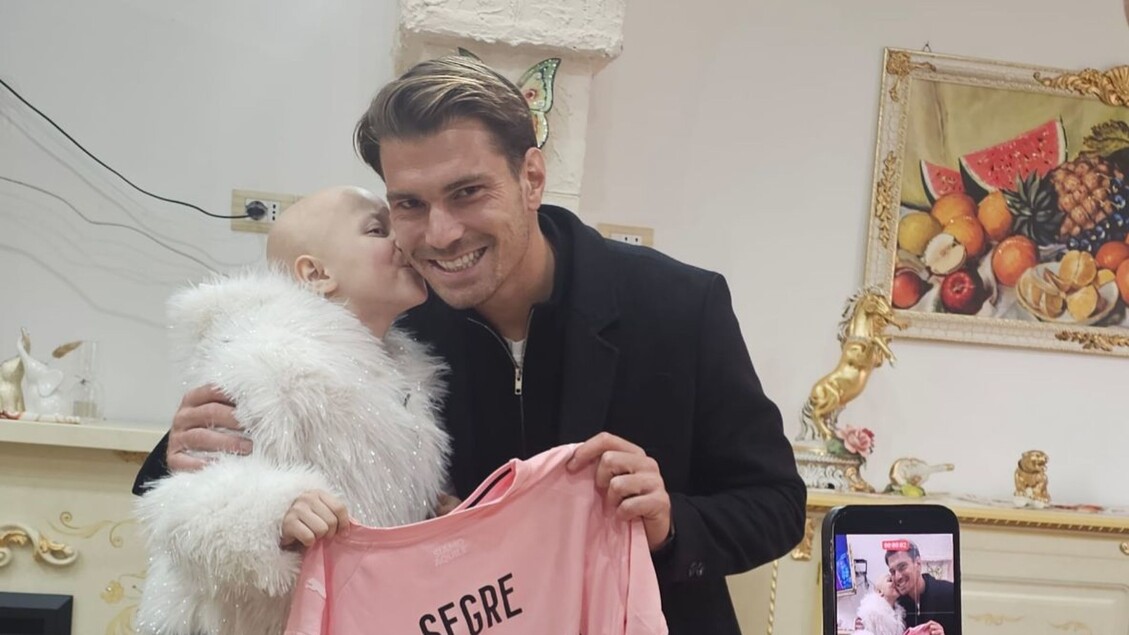 Morta Alessia La Rosa, la 'guerriera' tifosa del Palermo calcio, aveva otto anni. Un uomo tiene una maglietta rosa mentre un bambino lo bacia sulla guancia. Un'altra persona tiene un orsacchiotto bianco. La scena si svolge in un ambiente interno, con elementi di abbigliamento visibili.