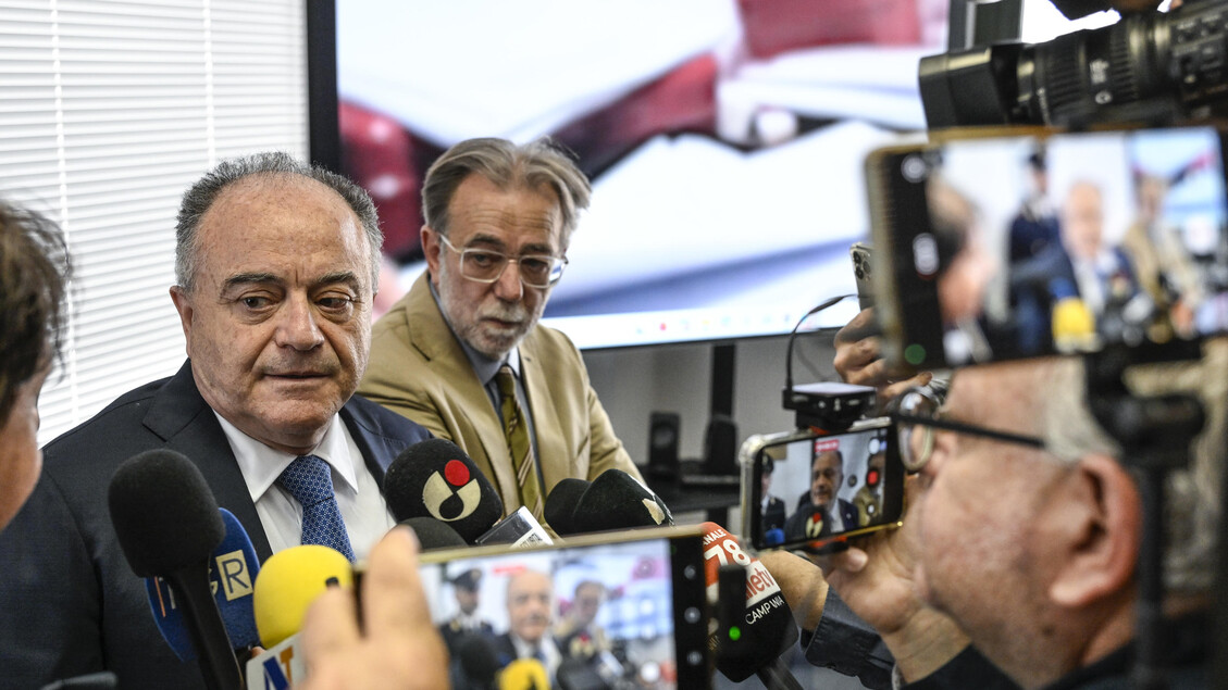 Durante una conferenza stampa, un gruppo di persone tiene in mano dei telefoni e parla con un uomo in giacca e cravatta. Un altro uomo, con un microfono, interagisce con i presenti. L'immagine mostra anche un operatore di telecamera e membri di una troupe televisiva. Il contesto riguarda la denuncia di Gratteri su dati rubati e venduti da poliziotti infedeli riguardanti calciatori e cantanti.