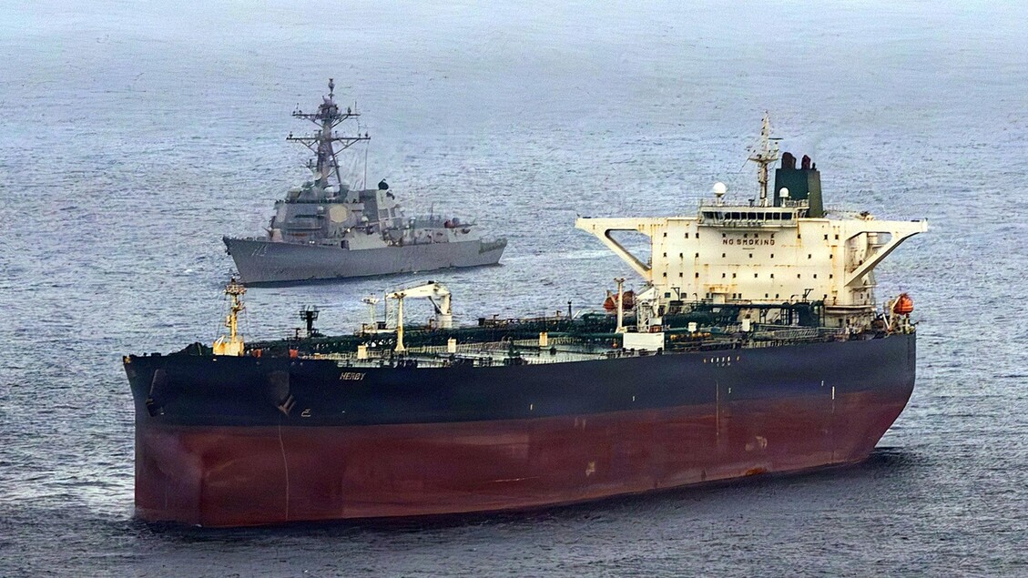 L'immagine mostra una grande nave in acqua, identificata come una nave mercantile. Il contesto &egrave; legato a tensioni geopolitiche tra Stati Uniti, Iran e Israele, suggerendo un ambiente di conflitto. La scena &egrave; all'aperto e rappresenta un elemento di trasporto marittimo.