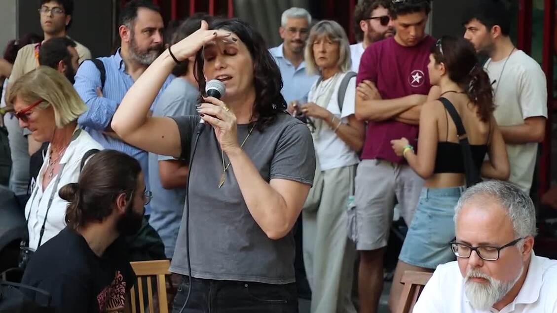Un'attivista donna tiene un microfono di fronte a una folla. Il contesto riguarda il sequestro di Spin Time, con gli attivisti che segnalano la presenza di bambini e anziani in strada, affrontando caldo e disagi. La scena evidenzia l'interazione tra la donna e il pubblico, sottolineando una situazione di protesta o sensibilizzazione.
