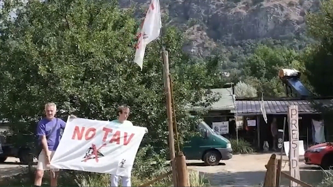 Un militante no Tav si trova in Val di Susa, esprimendo il suo amore per il territorio e la sua opposizione alla devastazione. Un uomo tiene un cartello con la scritta NO TAV. Sono visibili anche un edificio con segnali e un albero. L'ambiente &egrave; all'aperto.