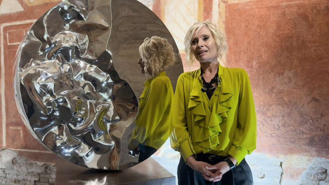 Una donna in una camicia gialla si trova accanto a una grande scultura d'argento. Il progetto si intitola Lumen ex terra ed &egrave; presentato alle Case Romane del Celio, realizzato da Helidon Xhixha. La scultura &egrave; parte di un evento artistico.