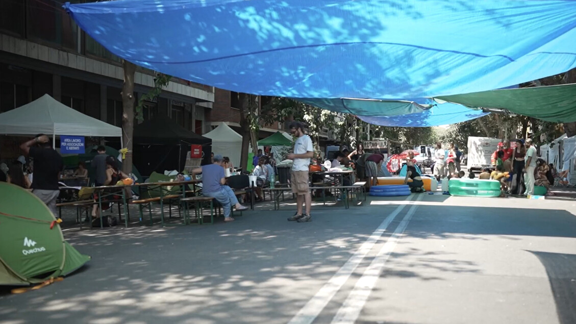 Un gruppo di persone &egrave; seduto per strada sotto un telo blu. La situazione &egrave; descritta come triste e preoccupante riguardo a un bene comune, secondo le parole di Fratoianni. L'ambiente &egrave; urbano, caratterizzato da un mercato e da spazi pubblici all'aperto. &Egrave; presente anche una tenda che fornisce ombra.