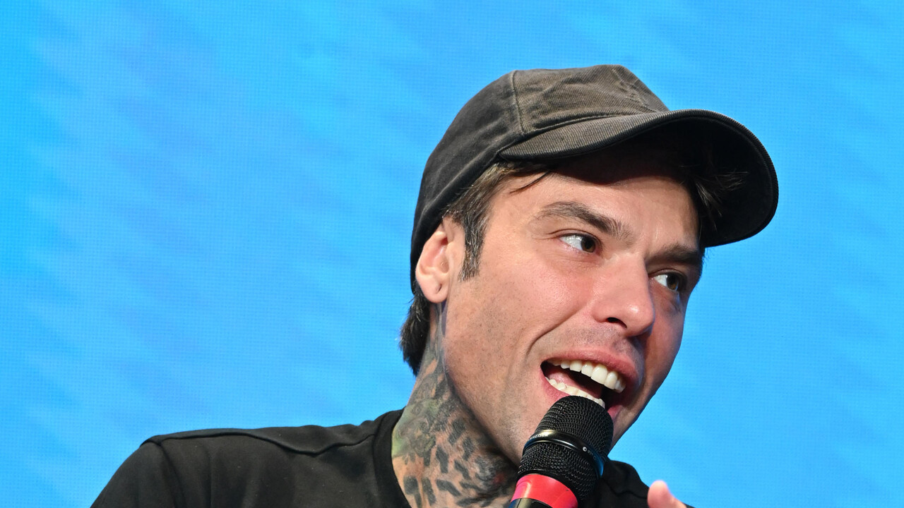 Fedez &egrave; riconoscibile nell'immagine. Si presenta con tatuaggi su braccia e viso, mentre tiene un microfono. Indossa un cappello e abbigliamento casual. L'ambientazione suggerisce un evento dal vivo, come un concerto.