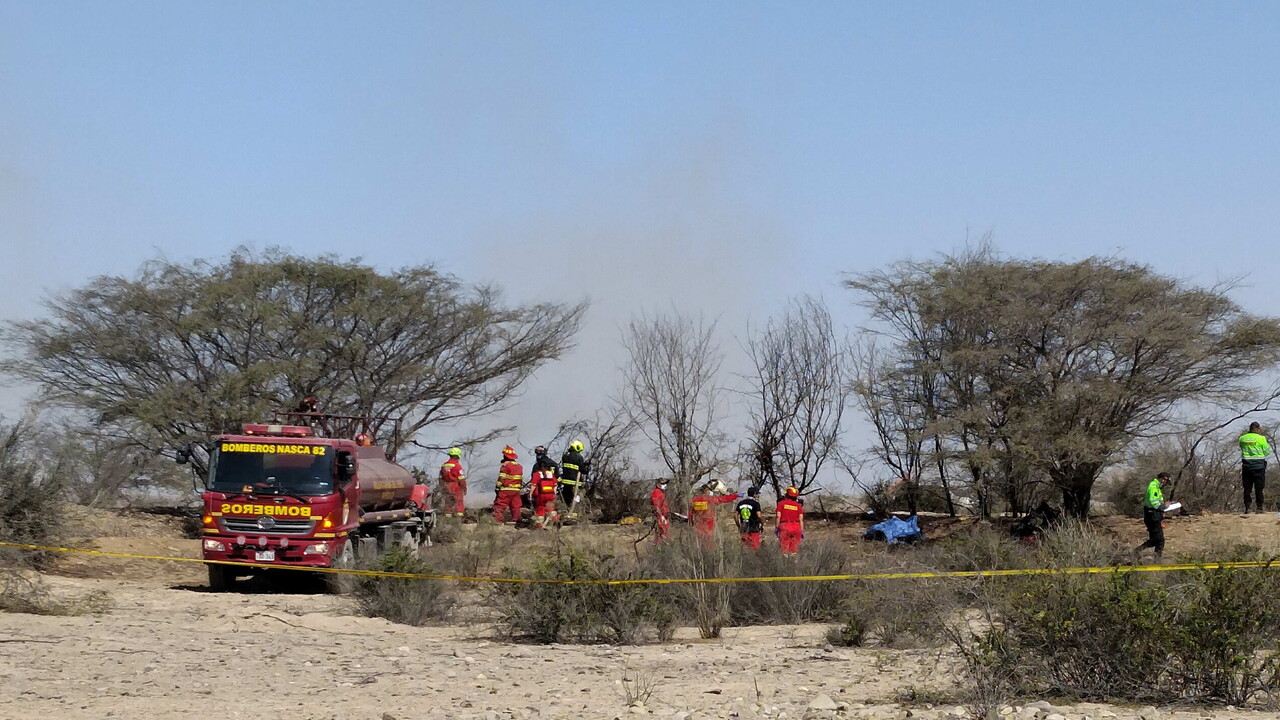 Un incidente aereo in Per&ugrave; ha causato la morte di 13 persone, tra cui 7 italiani. Nella scena, si osservano gruppi di persone in uniformi rosse e arancioni, probabilmente soccorritori o membri delle forze di emergenza. Sono presenti veicoli, tra cui un camion dei pompieri, in un contesto all'aperto con alberi e cielo visibile.