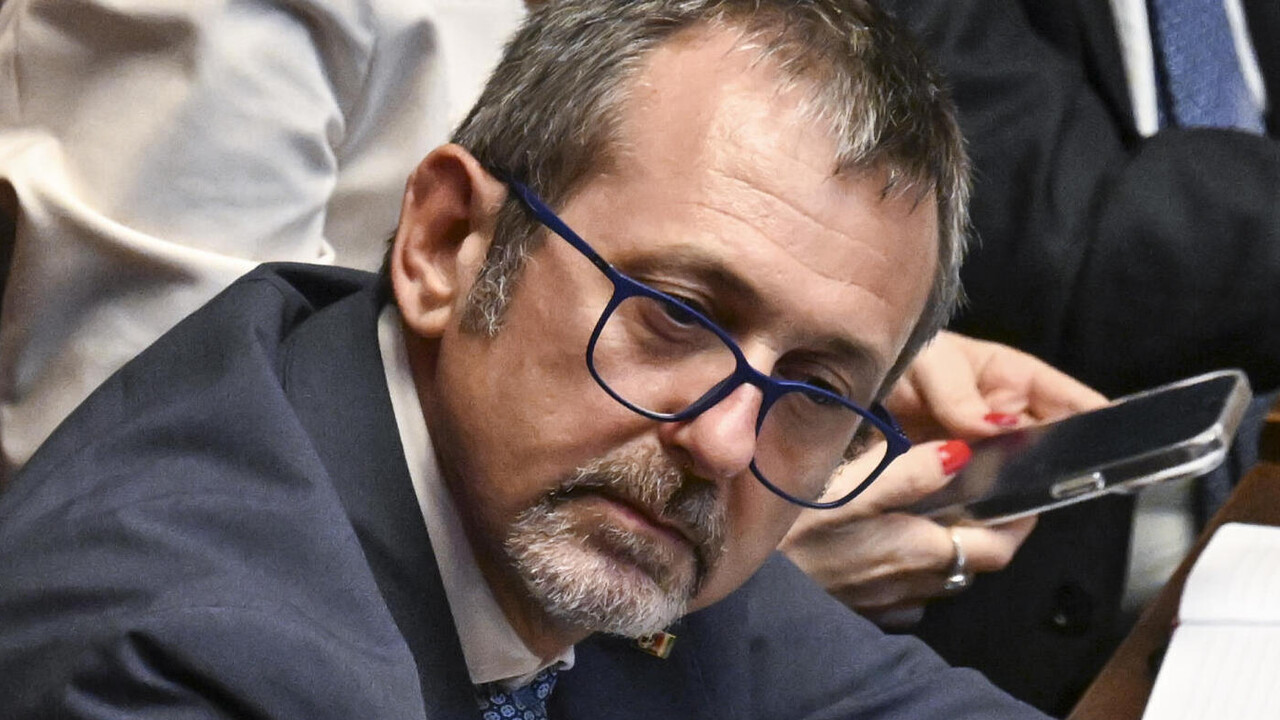 Andrea Delmastro &egrave; un uomo in giacca e cravatta seduto a una scrivania con un microfono. L'immagine si svolge in un ambiente interno.