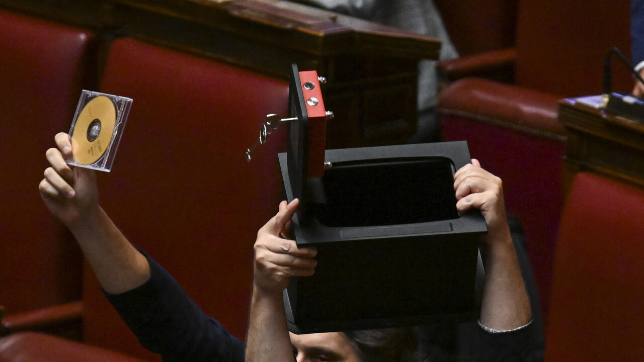 Un uomo tiene in mano una scatola e un CD. Il contesto mostra membri del M5S in Aula con striscioni che dicono fuori le chat e aprite la cassaforte. Sono presenti altre persone, creando un'atmosfera di partecipazione. L'ambiente &egrave; interno e include elementi di arredamento.