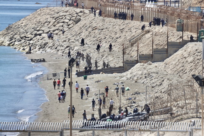 Un gruppo di persone cammina su una spiaggia sabbiosa a Ceuta. L'ambiente &egrave; all'aperto, con acqua e natura visibili. La scena si svolge lungo la costa, vicino alla linea di riva. L'Italia propone hub per migranti simili a quelli dell'Albania in un incontro post-Ceuta.