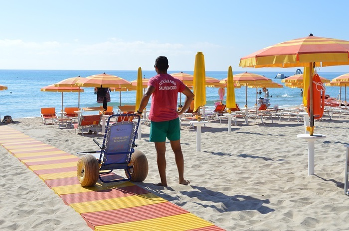 Un uomo spinge una sedia a rotelle su una spiaggia. La scena si svolge all'aperto, con il mare e la sabbia visibili. &Egrave; estate e il cielo &egrave; sereno. L'uomo &egrave; un adulto di sesso maschile. La spiaggia &egrave; un luogo di svago e natura, con acqua e terreno sabbioso.