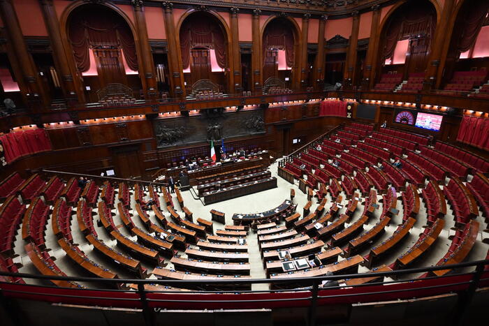 L'immagine rappresenta l'Aula della Camera, un ampio spazio interno con file di sedili e tavoli in legno. Si svolgono dichiarazioni di voto al decreto fiscale. L'ambiente &egrave; caratterizzato da un'architettura tipica di un edificio parlamentare, simile a un auditorium o a un teatro. Sono presenti persone che partecipano all'evento.
