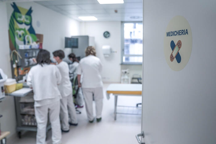 L'immagine mostra un reparto ospedaliero, con un gruppo di persone in una stanza. Tra di loro, si riconosce una persona che indossa pantaloni bianchi e un'altra figura sfocata con un camice bianco. L'ambiente &egrave; caratterizzato da elementi architettonici tipici di un ospedale, con attrezzature mediche e mobili presenti.