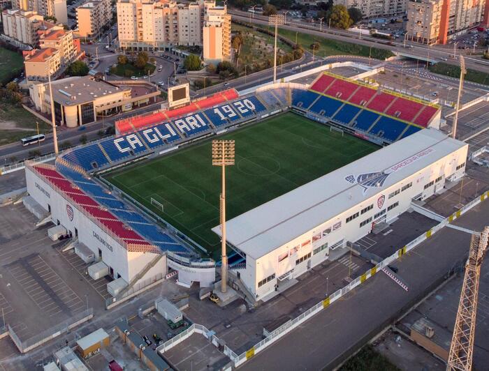 L'immagine mostra lo stadio Cagliari Unipol Domus, un grande stadio di calcio con un ampio campo verde. Sono presenti bandiere blu e rosse. Si osserva anche un'architettura bianca e un'area esterna. L'immagine include una vista aerea dello stadio, che &egrave; un'arena dedicata a eventi sportivi.