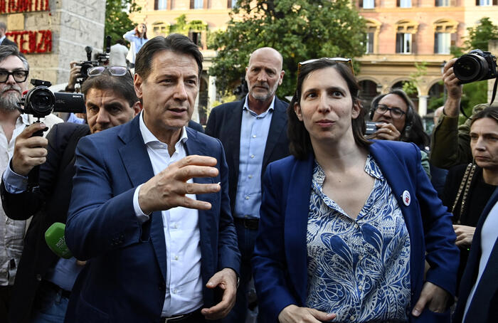 L'immagine rappresenta una maratona sui referendum, con i leader del Partito Democratico, del Movimento 5 Stelle, di Avs e di +E. Sono visibili un uomo e una donna, entrambi in abiti formali, con l'uomo che indossa un completo e la donna una giacca blu. L'ambientazione &egrave; all'aperto, e ci sono altre persone presenti. Un uomo tiene un telefono in mano.
