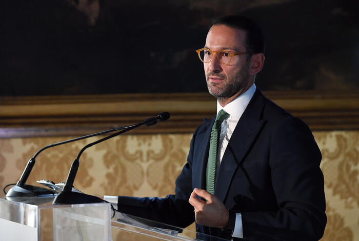 Un uomo in abito formale, con cravatta, si trova dietro a un podio. &Egrave; un oratore che sta parlando a un pubblico, composto da una folla di persone. Il contesto dell'immagine riguarda il rinvio della nomina del presidente della Consob.