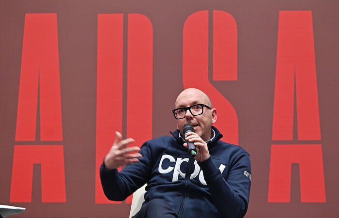 A Torino si svolge la presentazione di PHOTOANSA 2025. Un uomo &egrave; seduto su una sedia e tiene un microfono. Sono presenti altre persone e un pubblico.
