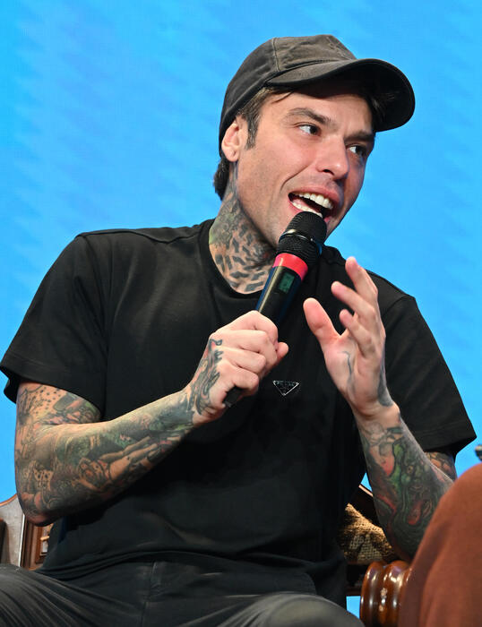 Fedez &egrave; riconoscibile nell'immagine. Si presenta con tatuaggi su braccia e viso, mentre tiene un microfono. Indossa un cappello e abbigliamento casual. L'ambientazione suggerisce un evento dal vivo, come un concerto.