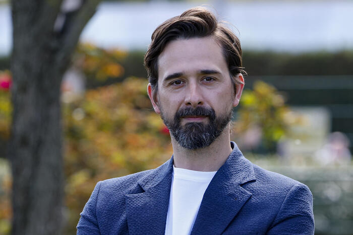 L'immagine mostra un uomo con barba e baffi, vestito con una giacca blu. Il suo volto &egrave; ben visibile, evidenziando i tratti distintivi. Il contesto &egrave; serio, poich&eacute; si fa riferimento a Davide Iacopini, che &egrave; in condizioni gravi, e a uno studente colpito. L'ambiente sembra essere all'aperto, con la presenza di alberi.