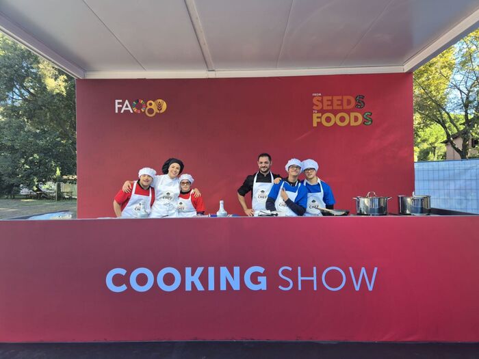 Un gruppo di persone indossa cappelli da chef e si trova dietro un tavolo rosso all'esposizione Fao per un concorso culinario dedicato all'inclusione. L'evento &egrave; caratterizzato da un'atmosfera di show culinario, con un focus su cibi e semi.