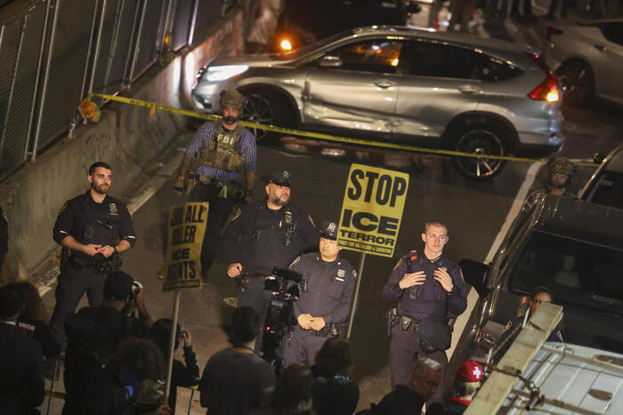 A New York, si verificano scontri dopo che l'ICE spara a un uomo. Sono presenti agenti di polizia in strada. Un'auto con un'ammaccatura si trova nella scena. Un uomo in uniforme nera tiene una macchina fotografica. La situazione &egrave; identificata come una scena del crimine.