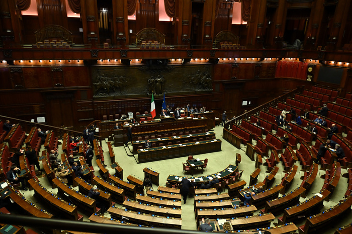 La scena si svolge in un'aula del Parlamento, caratterizzata da file di tavoli e sedie in legno. Si discute di una legge elettorale, con polemiche tra le opposizioni. Il Partito Democratico esprime preoccupazione per un possibile scontro istituzionale. Sono presenti anche 250 emendamenti in discussione. L'ambiente &egrave; interno e riflette un contesto di governo e architettura formale.