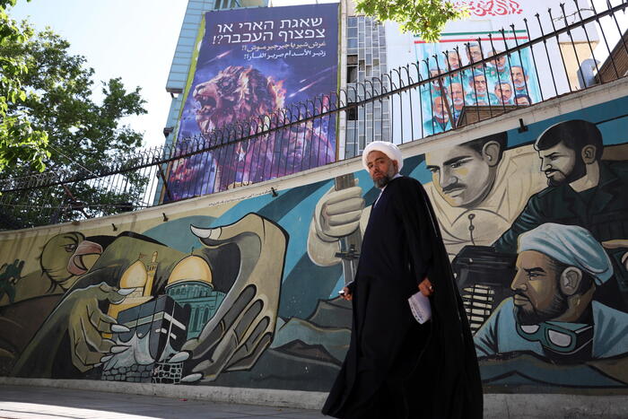 A Teheran, un uomo cammina davanti a un murale che rappresenta uccelli e un edificio. L'immagine riflette la vita quotidiana nella citt&agrave;, in un contesto di negoziazioni diplomatiche tra Stati Uniti e Iran. L'uomo indossa un abito nero e si trova in un ambiente all'aperto, caratterizzato da un murale artistico e alberi.