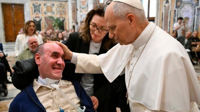 Aisla incontra il Papa e gli dona un ulivo. Il Papa, vestito con una tunica bianca, tocca la testa di un uomo. &Egrave; presente anche una donna in un abito nero. L'evento si svolge in un ambiente chiuso, probabilmente in un contesto religioso.