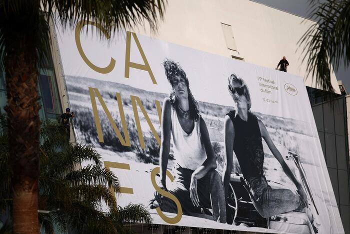 A Cannes, un grande cartellone pubblicitario &egrave; visibile su un edificio. Il cartellone &egrave; associato all'evento "Cannes 2026". Una persona &egrave; presente vicino al cartellone. L'ambiente &egrave; all'aperto, con alberi e palme nelle vicinanze.