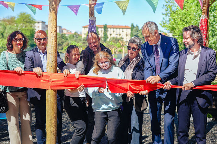 A Torino &egrave; stato inaugurato il nuovo orto di CasaOz. Un gruppo di persone tiene un nastro rosso, mentre un uomo in giacca e cravatta lo taglia. L'evento si svolge all'aperto, con alberi visibili sullo sfondo. Le persone indossano abbigliamento casual e formale.