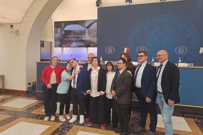 Un gruppo di ragazzi disabili, che include cuochi, meccanici e saldatori, partecipa a un progetto scuola-lavoro. Sono presenti insieme davanti a uno schermo e posano per una foto. Ludovico Einaudi &egrave; riconoscibile nell'immagine. Il contesto &egrave; indoor, con persone vestite in abiti formali.