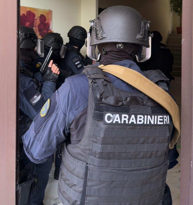 L'immagine mostra un gruppo di poliziotti in una porta, coinvolti in un'operazione legata a sequestri di persona, torture e bombe, con misure per sei persone. Sono presenti elementi di sicurezza come armature e caschi. Un testo riconosciuto menziona "CARABINIERI". L'atmosfera suggerisce un contesto di intervento delle forze dell'ordine.