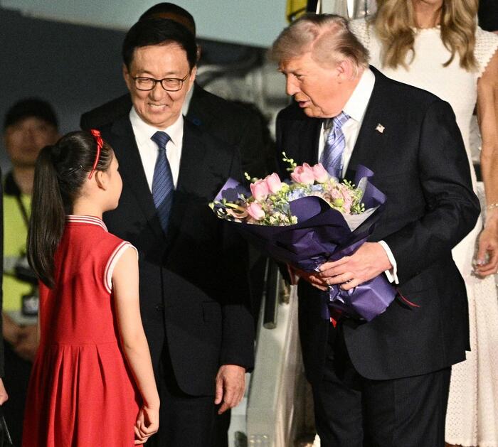 Un uomo in abito formale tiene un bouquet di fiori. &Egrave; presente anche una ragazza in un vestito rosso. Han Zheng e Donald Trump sono riconoscibili nell'immagine. L'ambientazione suggerisce un contesto di cerimonia.