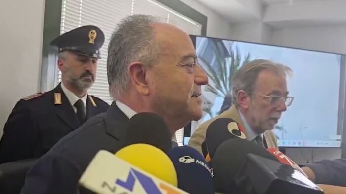 Durante una conferenza stampa, un gruppo di uomini in abiti formali parla in microfoni. Tra di loro, ci sono uomini in uniforme e in giacca. Gratteri afferma: "Rubati e venduti da poliziotti infedeli dati su calciatori e cantanti". La scena include anche una folla di persone, suggerendo un evento ufficiale.