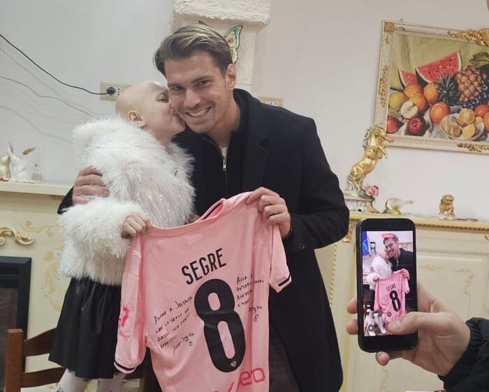 Morta Alessia La Rosa, la 'guerriera' tifosa del Palermo calcio, aveva otto anni. Un uomo tiene una maglietta rosa mentre un bambino lo bacia sulla guancia. Un'altra persona tiene un orsacchiotto bianco. La scena si svolge in un ambiente interno, con elementi di abbigliamento visibili.