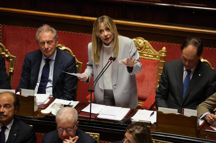 Giorgia Meloni &egrave; riconoscibile nell'immagine mentre parla a un podio con un microfono. Ci sono persone sedute sullo sfondo. L'evento si svolge in un contesto formale, probabilmente legato a questioni governative. La scena include un pubblico e rappresenta un momento di discussione su interessi nazionali.