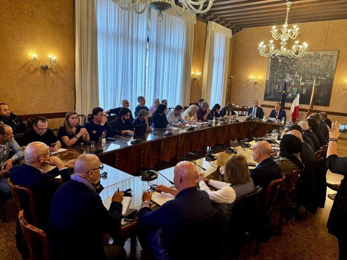 Un gruppo di persone &egrave; seduto attorno a un lungo tavolo in una sala conferenze. Un uomo in giacca &egrave; presente e tiene un microfono. L'ambiente &egrave; interno e sembra essere un seminario o una riunione. La scena include anche sedie e un'illuminazione da lampadario.
