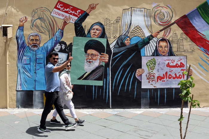 Un murale contro Stati Uniti e Israele si trova a Teheran. Davanti al murale, un uomo e una donna camminano. Il murale rappresenta donne che tengono una bandiera. Ali Khamenei &egrave; riconoscibile nell'immagine. La scena include persone in un contesto di protesta, con elementi artistici e urbani.