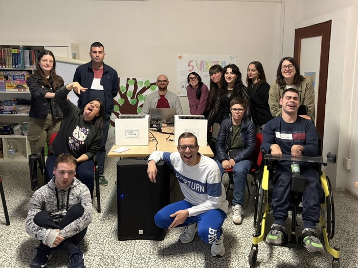 Un laboratorio radiofonico inclusivo per ragazzi con disabilit&agrave; si trova a Chieti. Un gruppo di persone posa per una foto. Tra di loro, ci sono persone sedute su sedie e una persona in sedia a rotelle. L'ambiente &egrave; interno e presenta elementi di arredamento come sedie.