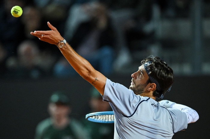 Lorenzo Musetti e Darderi avanzano al terzo turno degli Internazionali. Un uomo tiene una racchetta da tennis. L'immagine rappresenta un evento sportivo di tennis, con un pallone da tennis visibile.