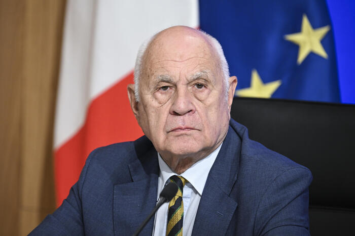 L'immagine ritrae un uomo in abito formale, con giacca e cravatta. &Egrave; un uomo adulto, identificato come un uomo d'affari. Il contesto suggerisce un ambiente ufficiale, probabilmente legato a un evento o a una situazione formale. La presenza di altre persone indica che si tratta di un contesto sociale o professionale.