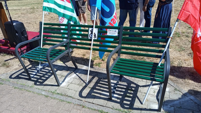 Nel Parco Carr&agrave; di Alessandria &egrave; inaugurata la Panchina Inclusiva. Un gruppo di persone si trova accanto a una panchina verde con gambe in metallo. La scena &egrave; all'aperto, su una superficie di mattoni, circondata da erba e piante.