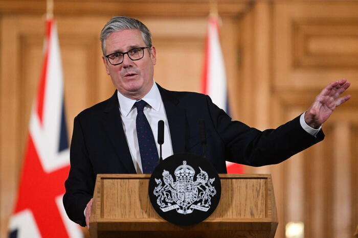 Keir Starmer, il Primo Ministro britannico, tiene una conferenza stampa riguardo al divieto dei social media per i teenager. &Egrave; riconoscibile nella foto mentre sta in piedi a un podio, con una mano alzata. Indossa un abito formale con cravatta. Attorno a lui ci sono persone che compongono il pubblico, creando un'atmosfera di discussione ufficiale.