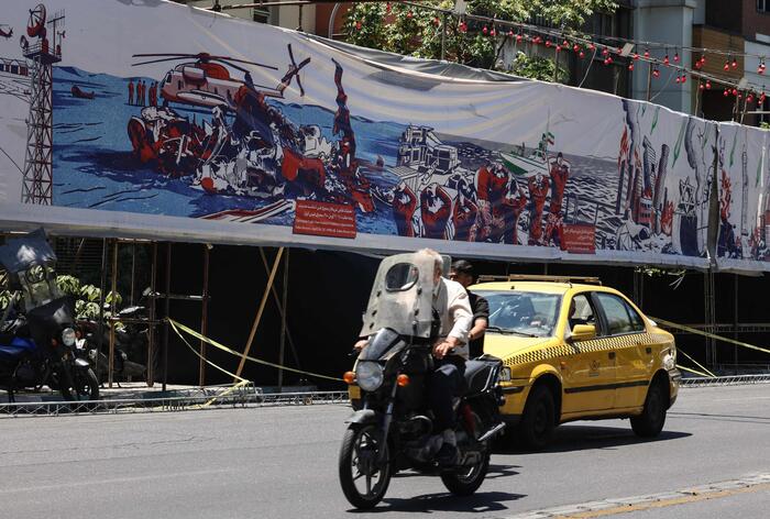 A Teheran, un uomo guida una motocicletta su una strada urbana. &Egrave; presente un banner con l'immagine di una nave. L'ambiente &egrave; caratterizzato da una strada con una recinzione e alberi. Sono visibili anche un'auto e altri veicoli. La scena riflette un contesto di conflitto tra Iran, Stati Uniti e Israele.