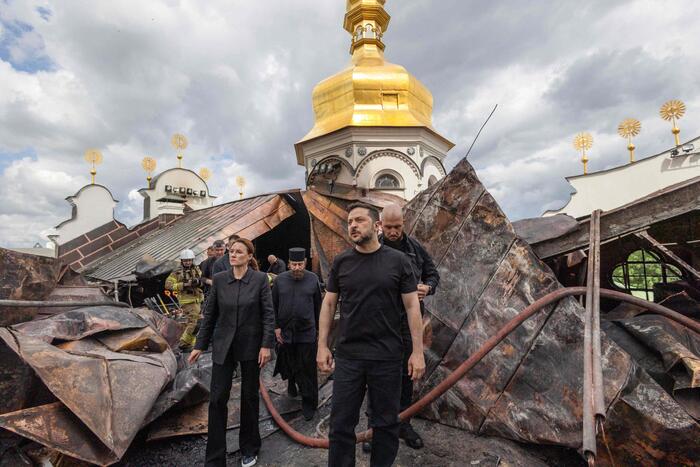 L'immagine mostra il presidente Zelensky sul luogo di un incendio, circondato da un gruppo di persone all'aperto. Si vede un edificio danneggiato, caratterizzato da un'architettura dorata e bianca con una torre. Il cielo &egrave; nuvoloso. Le persone indossano abbigliamento adatto per l'esterno.