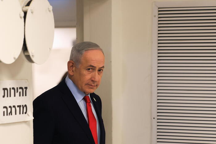 Nell'immagine, il Primo Ministro israeliano Benjamin Netanyahu &egrave; presente, indossando un abito formale con cravatta. Si osservano anche altre persone, suggerendo un contesto di celebrazione o vittoria, con riferimenti agli Stati Uniti e all'Iran. Un primo piano di una finestra &egrave; visibile, ma non &egrave; chiaro il suo significato nel contesto. La scena si svolge in un ambiente interno, probabilmente durante un evento ufficiale.